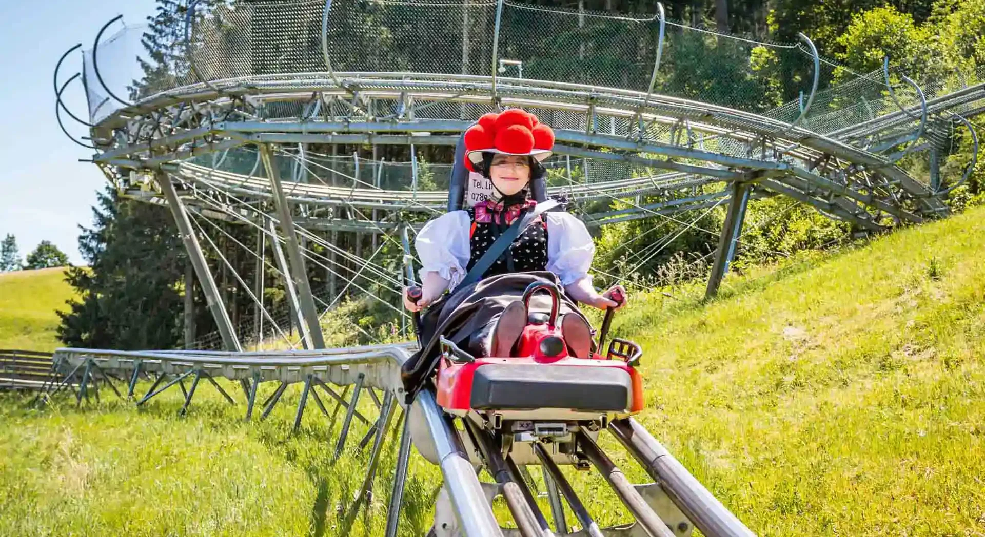 Eine Frau in traditioneller Tracht fährt auf der Sommerrodelbahn in Gutach durch die grüne Berglandschaft.